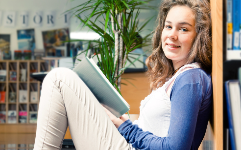 teen girl sitting against shelf in library reading a book