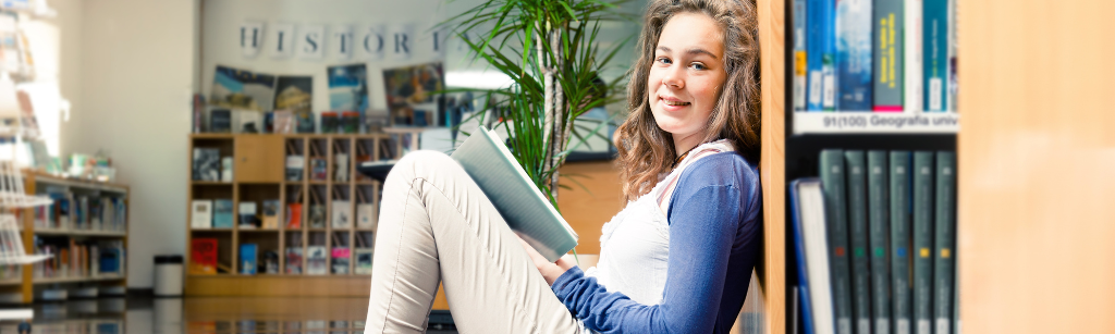 teen girl sitting against shelf in library reading a book