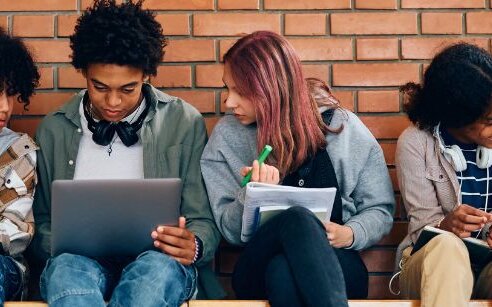 high school students sitting on bench against brick wall with lap tops, talking and taking notes