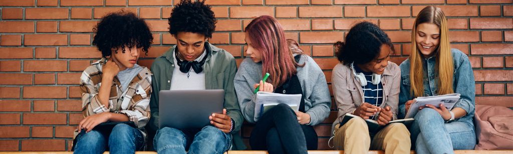 high school students sitting on bench against brick wall with lap tops, talking and taking notes
