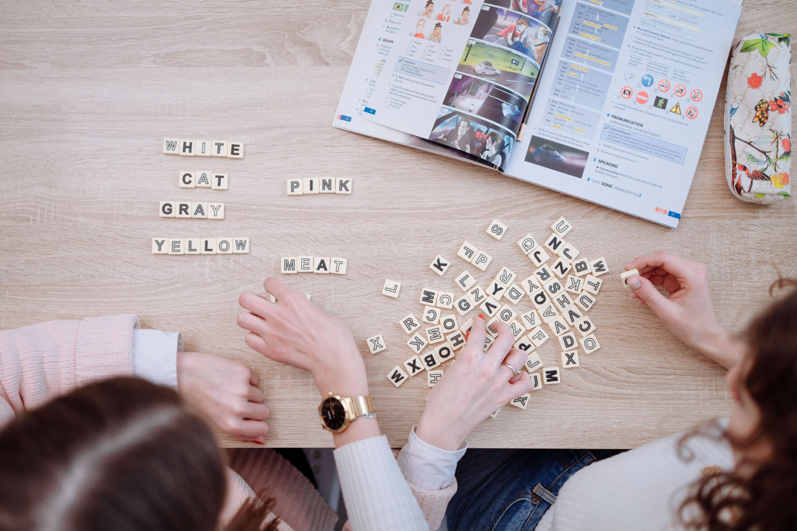 Overhead view of two people manipulating letter tiles to make words, with an instructional book in front of them.