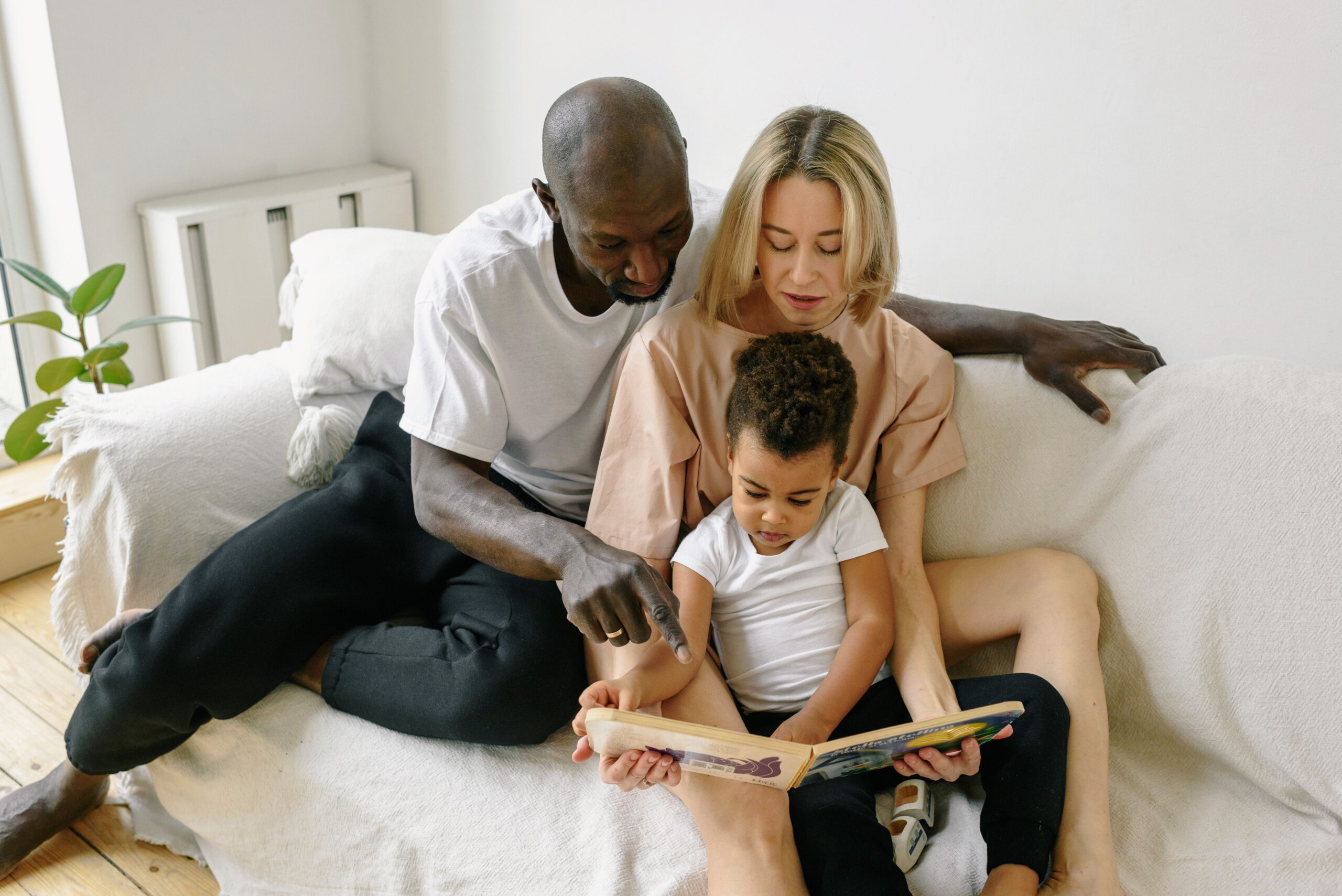 Multicultural family reading a picture book with young child on couch.
