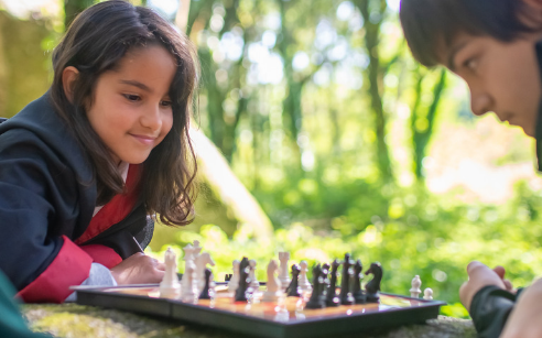 Smiling girl playing chess on a log with two boys in a forest.